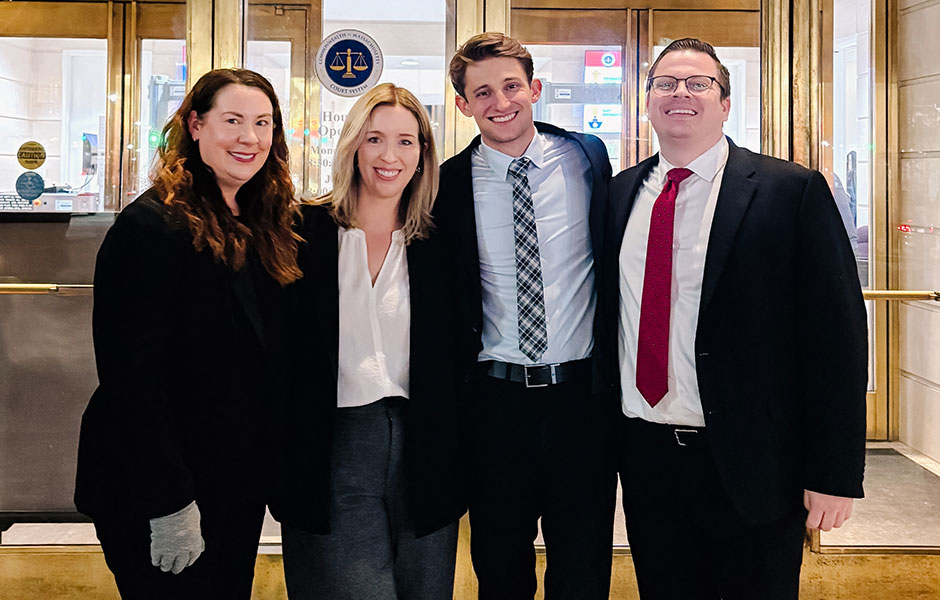 Four professionally dressed individuals, two women and two men, standing and smiling together in front of the glass doors of a Commonwealth of Massachusetts Court System building.